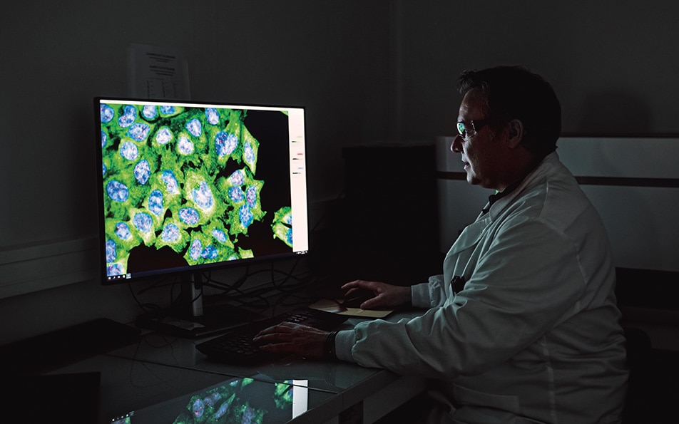 Un chercheur en blouse blanche regardant un écran affichant des images microscopiques de cellules colorées en vert et bleu sur un grand écran