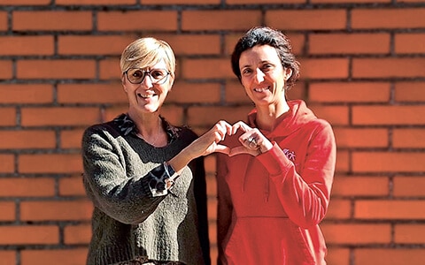 Deux femmes souriantes, l'une portant un pull vert et des lunettes, l'autre portant un sweat-shirt rouge, formant un cœur avec leurs mains devant un mur de briques.
