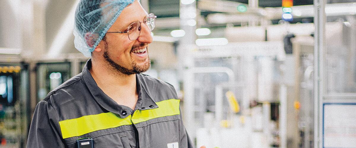 Un employé portant une charlotte bleue et des lunettes de sécurité, tenant un produit dans l'usine de Soual, entouré de machines industrielles.