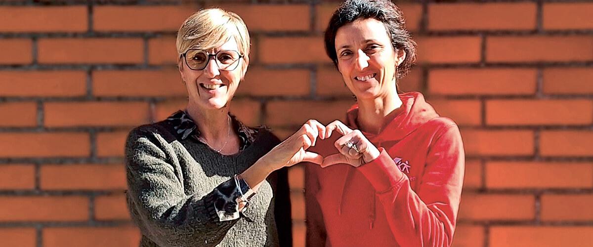 Deux femmes souriantes, l'une portant un pull vert et des lunettes, l'autre portant un sweat-shirt rouge, formant un cœur avec leurs mains devant un mur de briques.