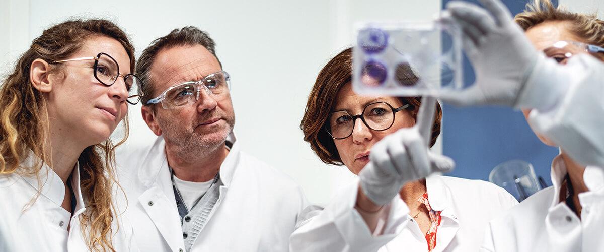 Quatre chercheurs en blouses blanches et lunettes de sécurité examinent attentivement une tablette avec des résultats expérimentaux.