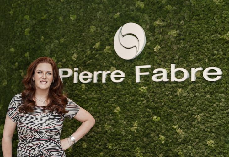 Imagem da Danielle Bibas na frente de uma parede verde de plantas verdes onde encontra-se a logo da Pierre Fabre e o nome da empresa em branco
