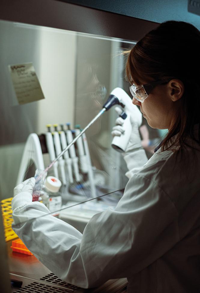Une chercheuse en blouse blanche et lunettes de sécurité regardant devantelle, avec un équipement de laboratoire en arrière-plan