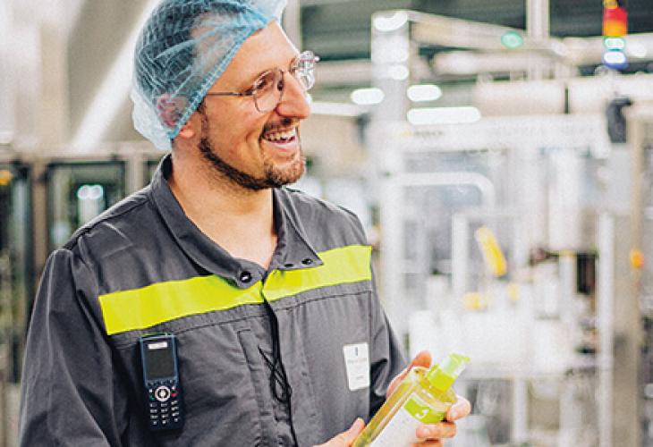 Un employé portant une charlotte bleue et des lunettes de sécurité, tenant un produit dans l'usine de Soual, entouré de machines industrielles.