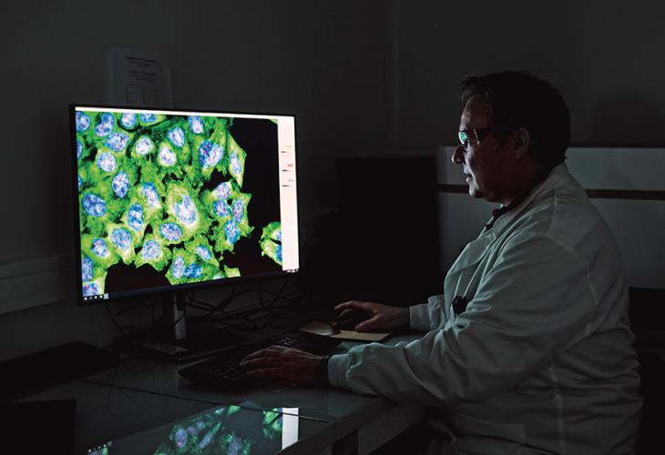 Un chercheur en blouse blanche regardant un écran affichant des images microscopiques de cellules colorées en vert et bleu sur un grand écran