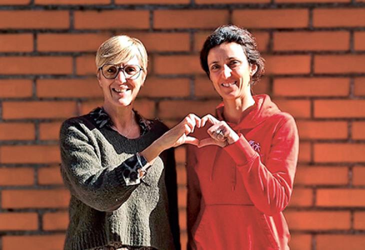 Deux femmes souriantes, l'une portant un pull vert et des lunettes, l'autre portant un sweat-shirt rouge, formant un cœur avec leurs mains devant un mur de briques.
